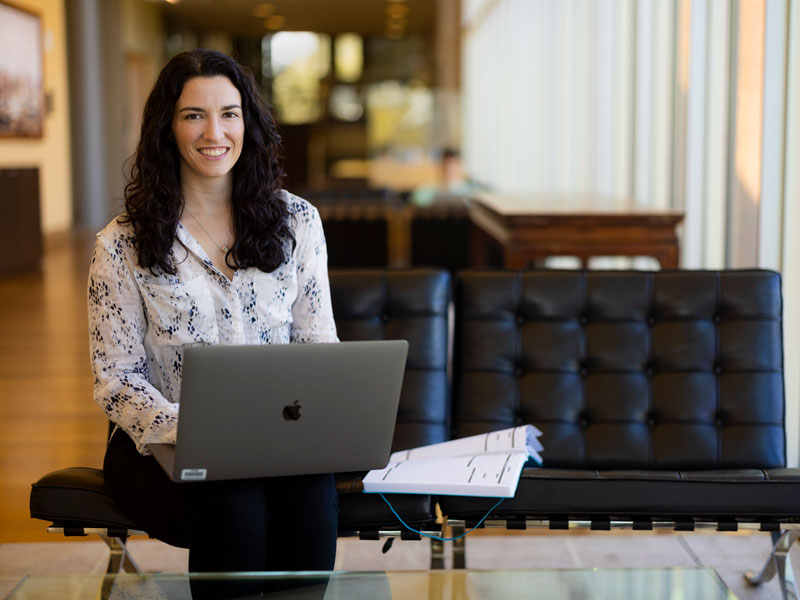 Young woman with long dark hair smiles while working on a laptop on a modern black couch.