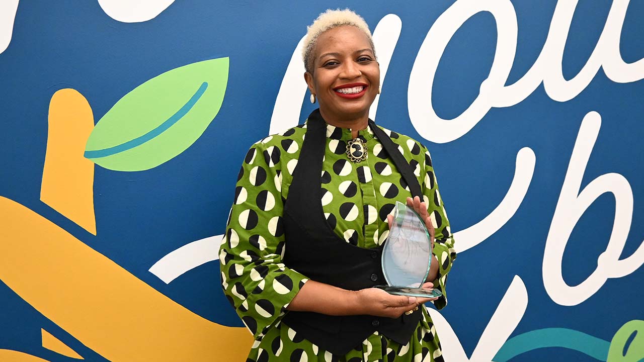 Woman in a green patterned dress smiles holding a glass award.