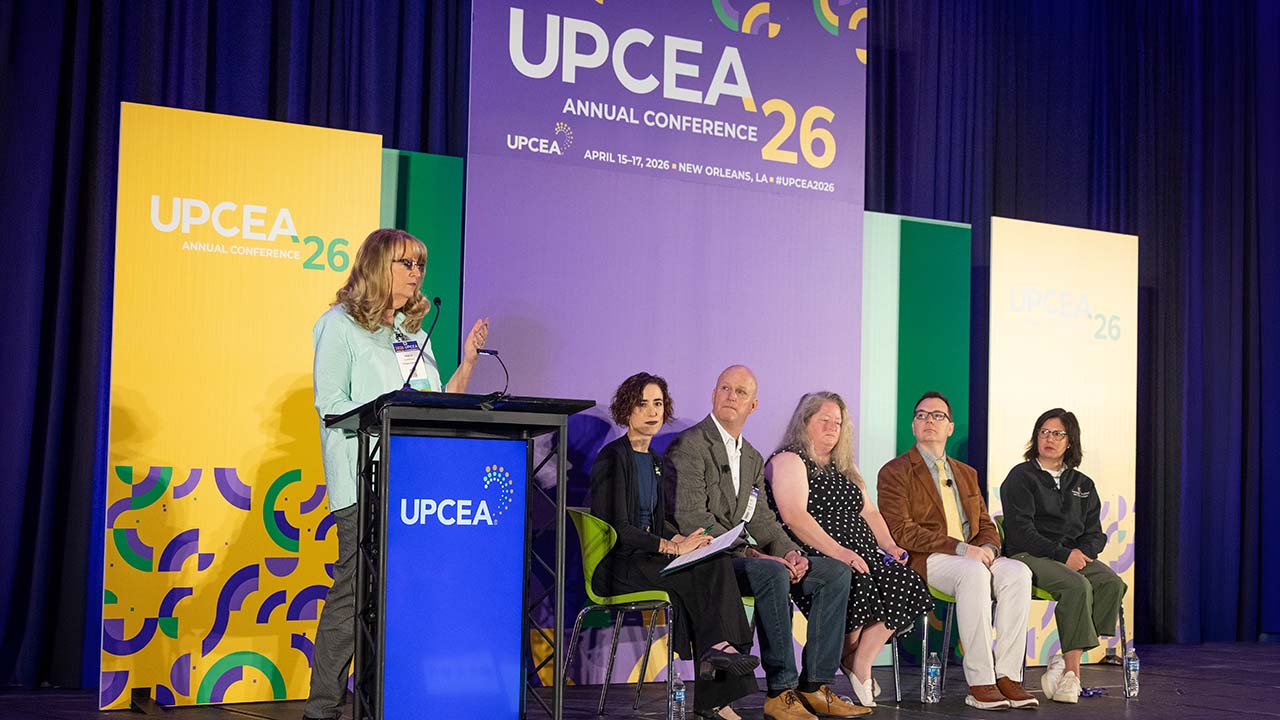 Woman speaks at podium before a panel of five people on a stage with UPCEA 26th conference banners.