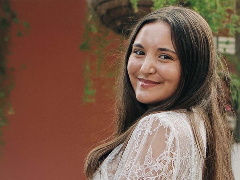 Young woman with long brown hair smiles over her shoulder outdoors.