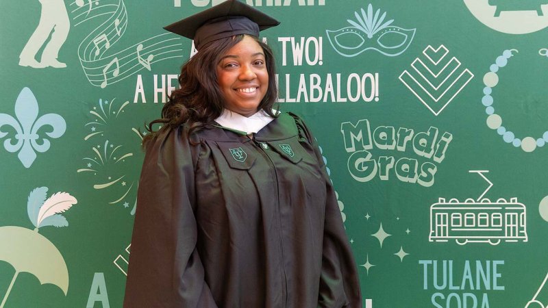 A graduate in a black cap and gown smiles broadly in front of a green Mardi Gras themed backdrop.