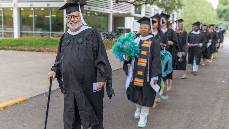 Graduates in caps and gowns walk in a procession, one with a cane, another with a bouquet.