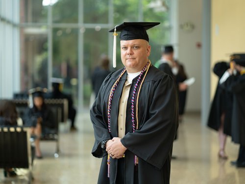 Man in graduation cap and gown stands with hands clasped.