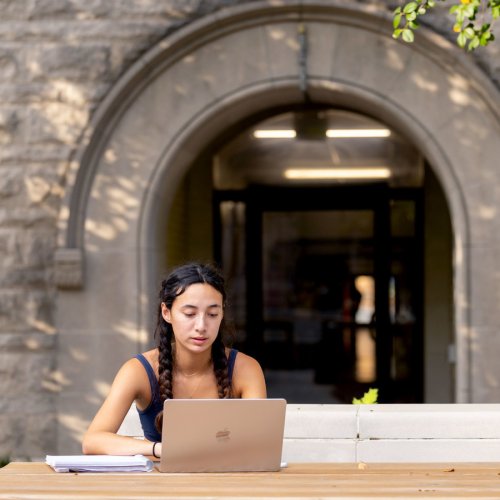 A young woman with braided hair uses a laptop at a picnic table outside a stone building.