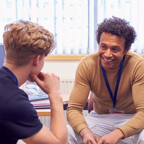A smiling man in a tan sweater talks to a young person in a classroom.