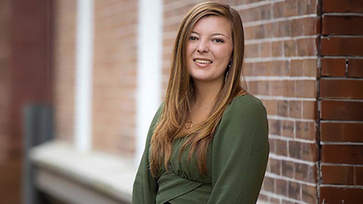 Young woman with long brown hair smiles, leaning against a brick wall.