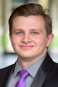 Young man wearing a black suit, gray shirt, and purple tie, smiling.