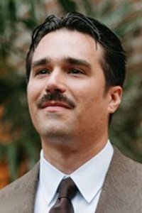Man with mustache in a brown suit and tie, looking up and to the left.