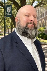 Bald man with a beard in a suit, smiling, with a Tulane banner and building behind him.