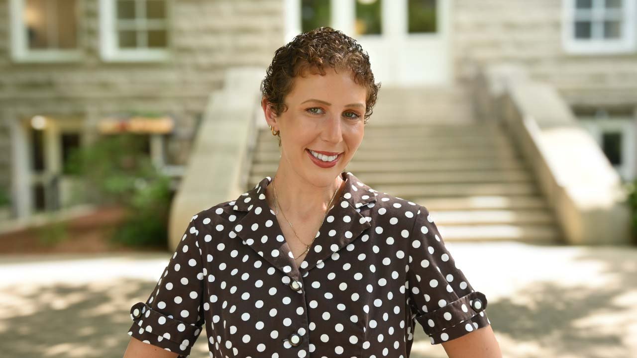 Woman with short curly hair smiles, wearing a brown polka-dot shirt, standing in front of a building.