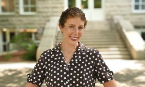 Woman with short curly hair smiles, wearing a brown polka-dot shirt, standing in front of a building.