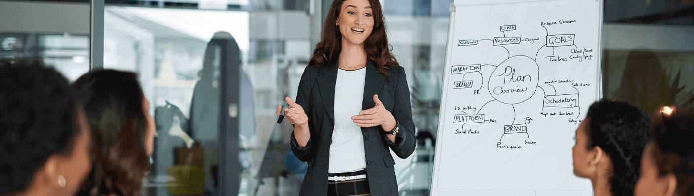 A woman giving a presentation to a conference room of people in front of a diagram