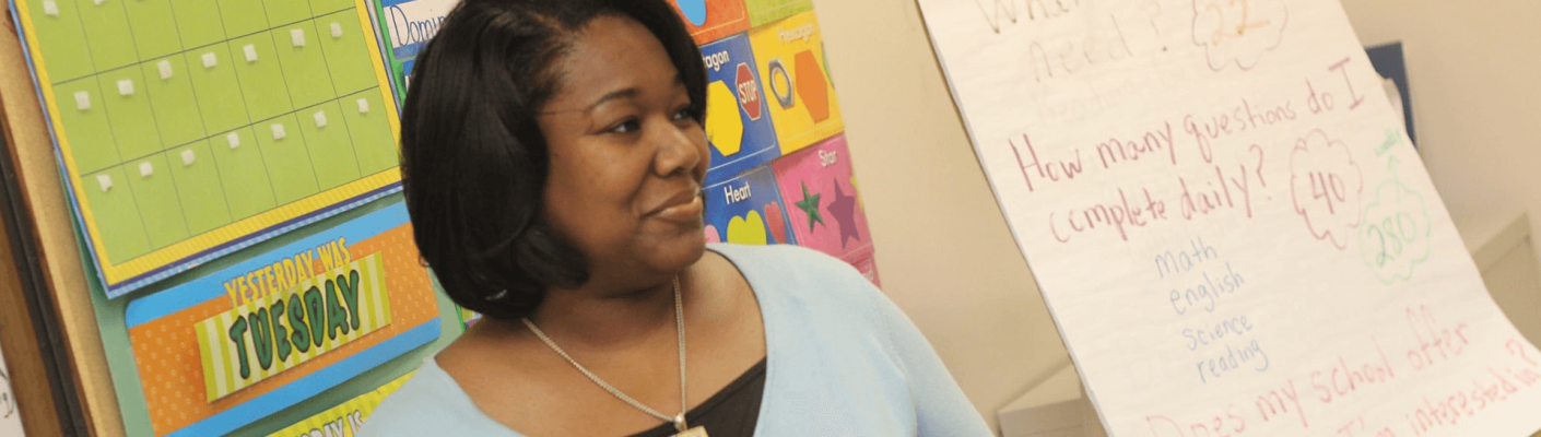 A teacher smiling next to education posters in a classroom
