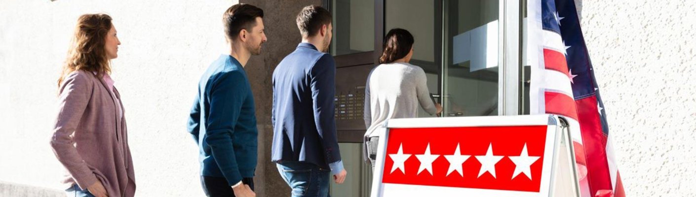 People line up to enter a building next to a sign reading “Vote Here” and a United States flag.