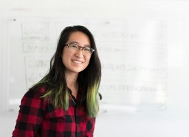 Young woman with green-streaked hair, wearing glasses and a red plaid shirt, smiling.